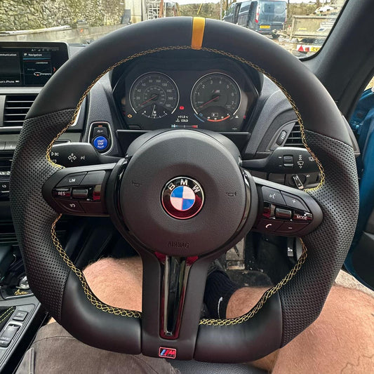 A custom black leather steering wheel with gold contrast stitching and a central BMW logo, installed in a car interior. The text 'Custom steering wheel' is overlaid at the top.